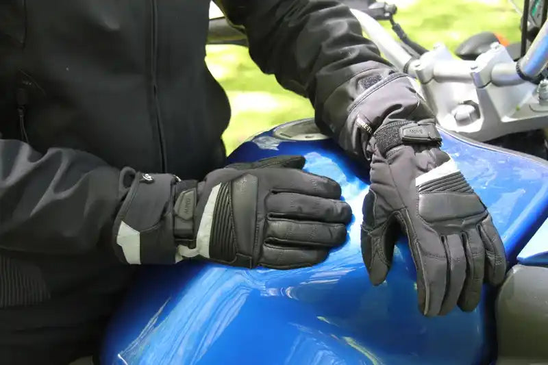 Black motorcycle riding gloves with gray and white reflective accents, worn by a rider sitting on a blue motorcycle.