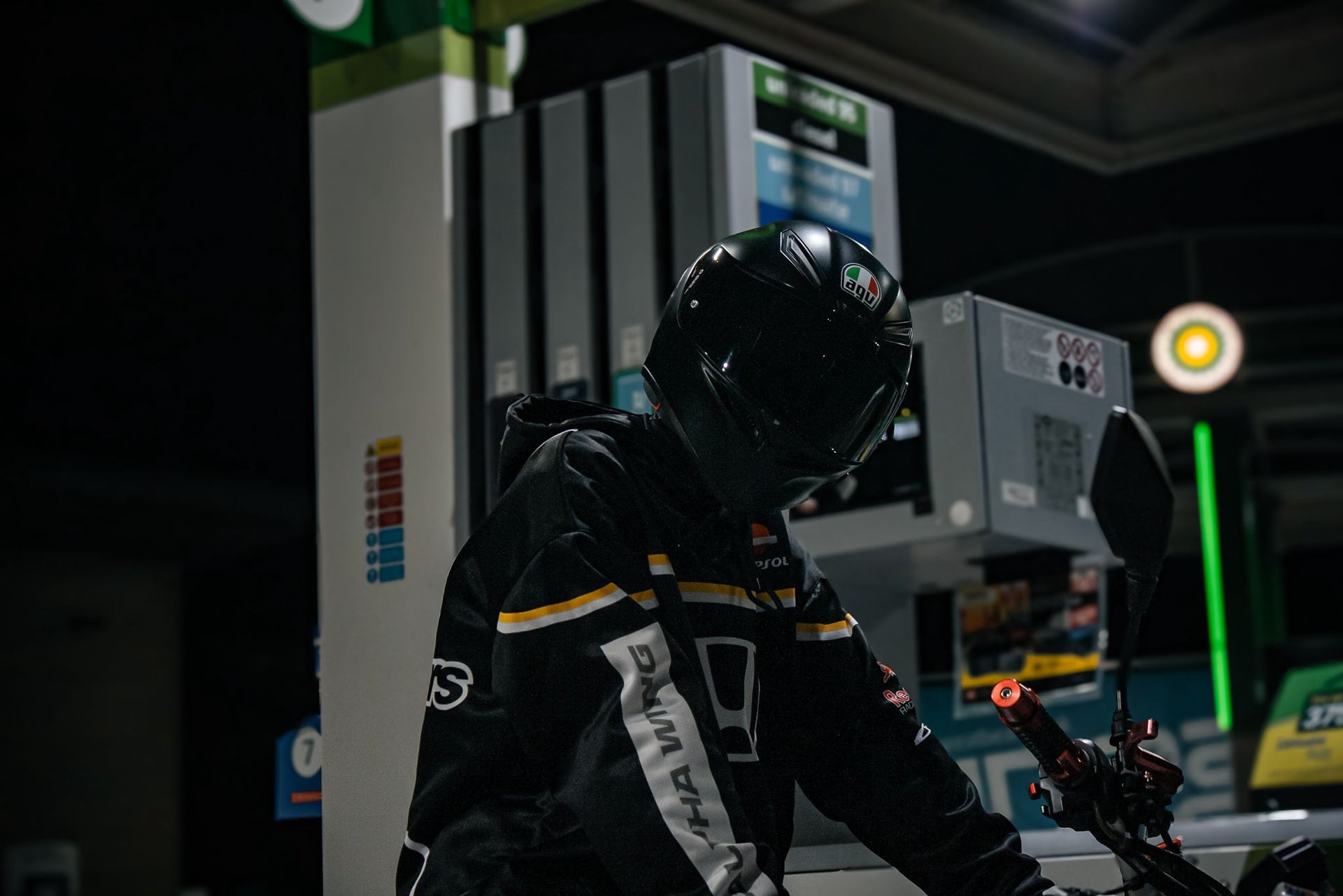 A person in a black full-face helmet and black riding armored hoodie with white and yellow stripes stands at a gas station pump at night, with a alpha wing brand name on his arm