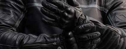 Black leather motorcycle gloves worn on a person's hands, shown against a dark leather jacket background.