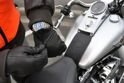 Black leather custom motorcycle gloves worn by a rider sitting on a silver motorcycle, with one glove being unzipped to reveal a watch inside.