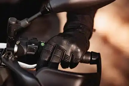 A close-up shot of a person's hand wearing a black leather motorcycle glove, gripping the handlebar of a bike.