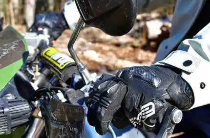 A close-up of a person wearing a black leather custom motorcycle glove with a white and gray sleeve, gripping the handlebar of a dirt bike outdoors in a wooded setting.