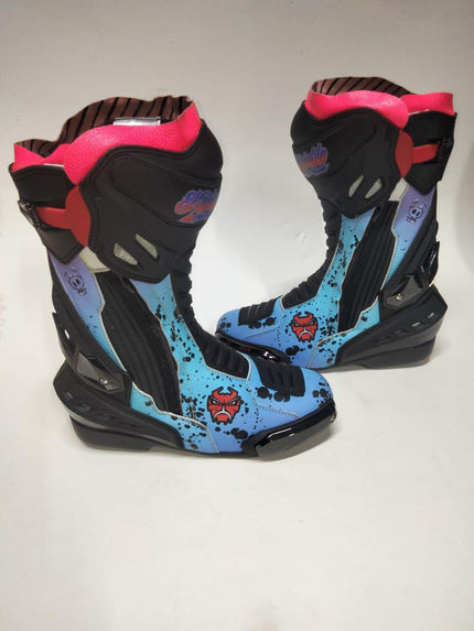 A pair of high-top motocross or racing boots with a black base, bright pink collar lining, and a light blue splatter-patterned side panel with red graphic accents.