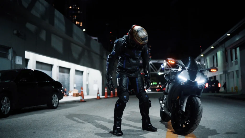 A motorcyclist in a black leather suit and helmet stands beside a sleek motorcycle on a dimly lit urban street at night.