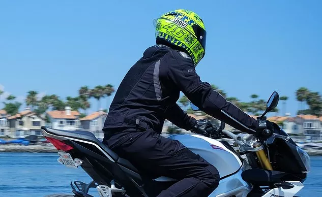 A motorcyclist in black gear and a bright yellow helmet rides along a waterfront with palm trees and homes in the background.