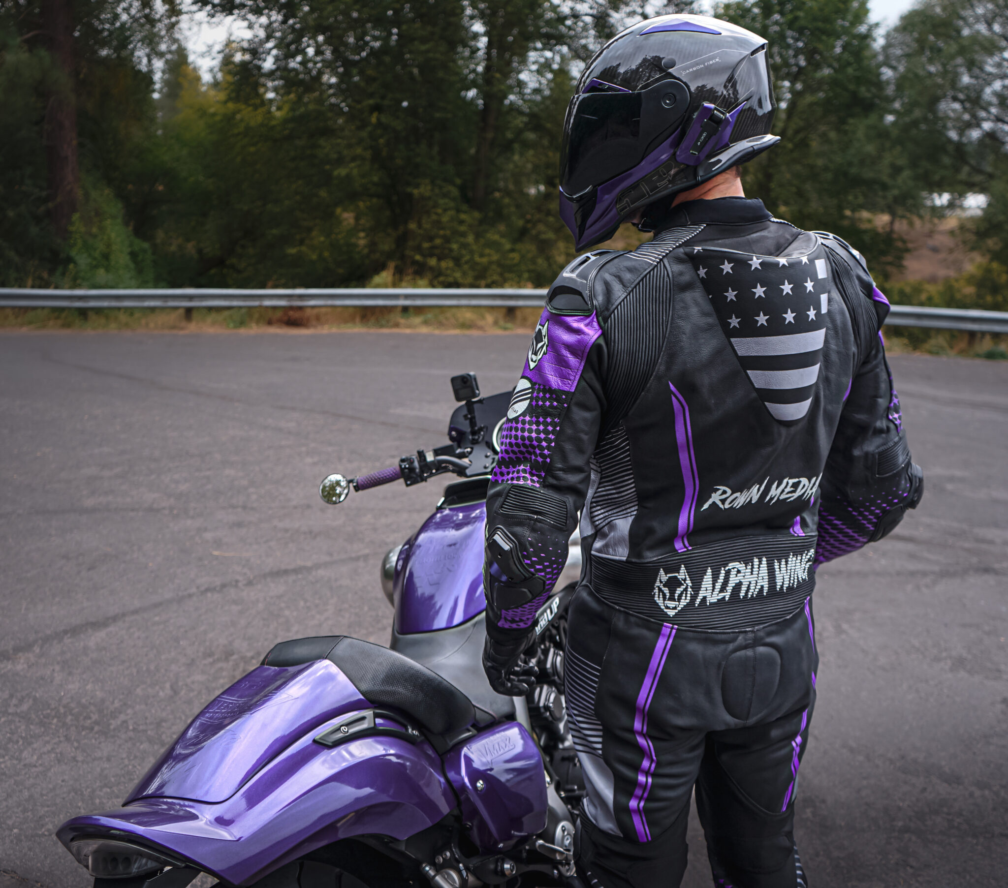 A person wearing a black and purple motorcycle suit and helmet stands next to a matching purple motorcycle, facing away toward a wooded area and a road with a metal guardrail.