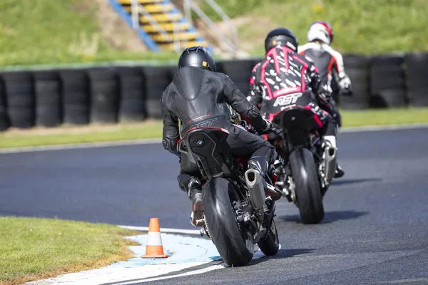 Three motorcycle riders in riding gear navigate a track, with a traffic cone marking the curve in the foreground.