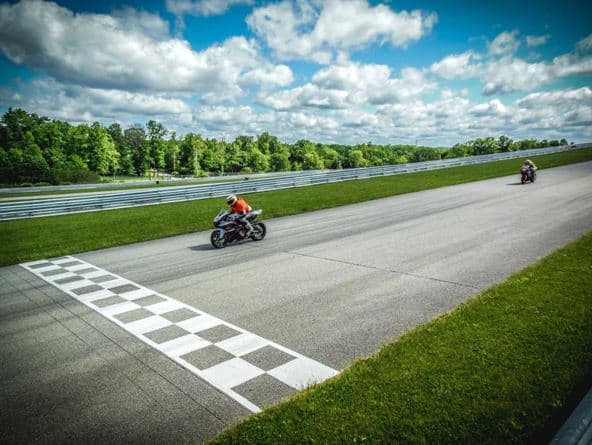 Two motorcyclists race near the finish line on a sunny day at a racetrack, surrounded by green trees and a blue sky.