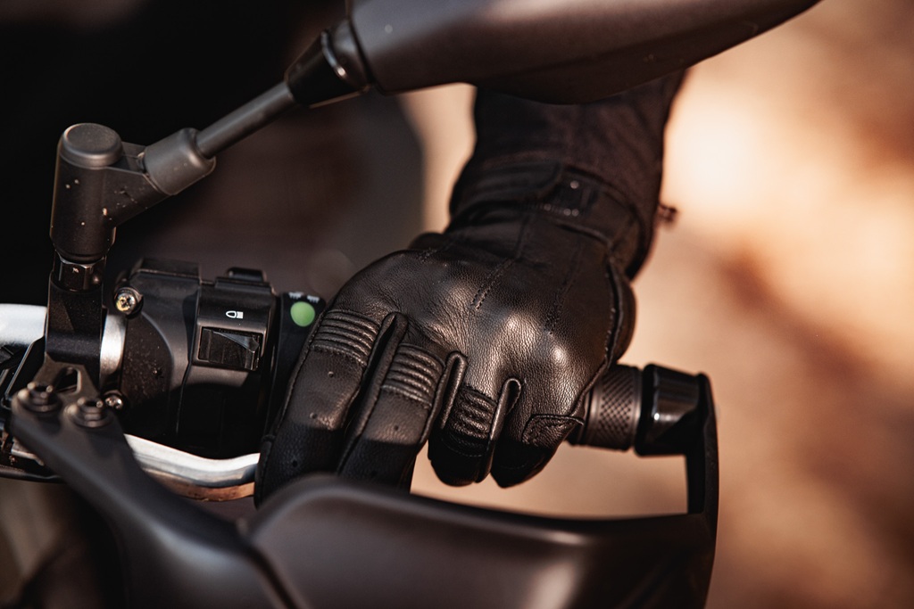 Close-up of a gloved hand gripping a motorcycle handlebar with control buttons and a lever, set in a blurred outdoor background.