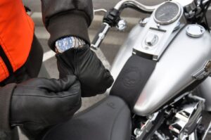 A person in a bright orange jacket adjusts a black leather glove while seated on a silver motorcycle, showcasing a sleek watch.
