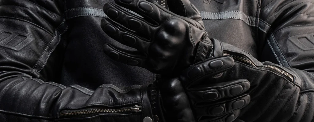 Close-up of a person wearing black leather motorcycle gloves, adjusting them with a textured leather jacket in the background.